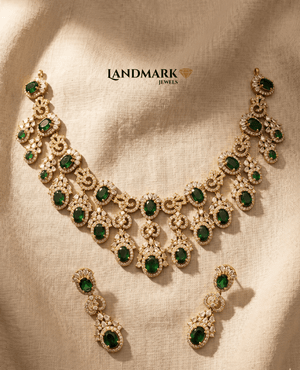 Ornate gold-plated necklace and earring set featuring oval emerald green stones and white sparkling crystals by Landmark Jewels, displayed on a textured fabric background.