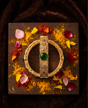 Antique gold-plated bangles with emerald green stones and floral Meenakari design on a traditional festive background.