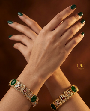 Antique gold-plated bangles with emerald green stones and floral Meenakari design on a traditional festive background.