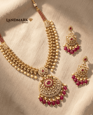 Landmark Jewels antique gold-plated necklace featuring ruby stones, pearl droplets, and matching heavy chandelier earrings on a beige fabric background.