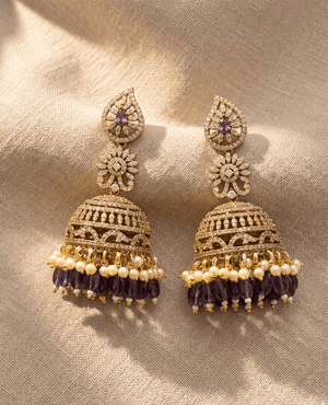 Grand tiered gold Jhumka earrings featuring a paisley top with a purple stone, an intensely detailed crystal dome, and layered fringes of white pearls and purple glass beads.