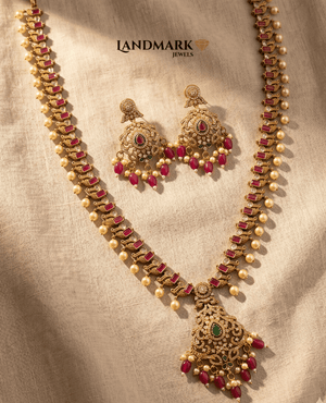 Landmark Jewels antique gold-plated long haram necklace and earring set featuring red ruby stones, green emerald accents, and hanging pearl drops on a beige fabric background.