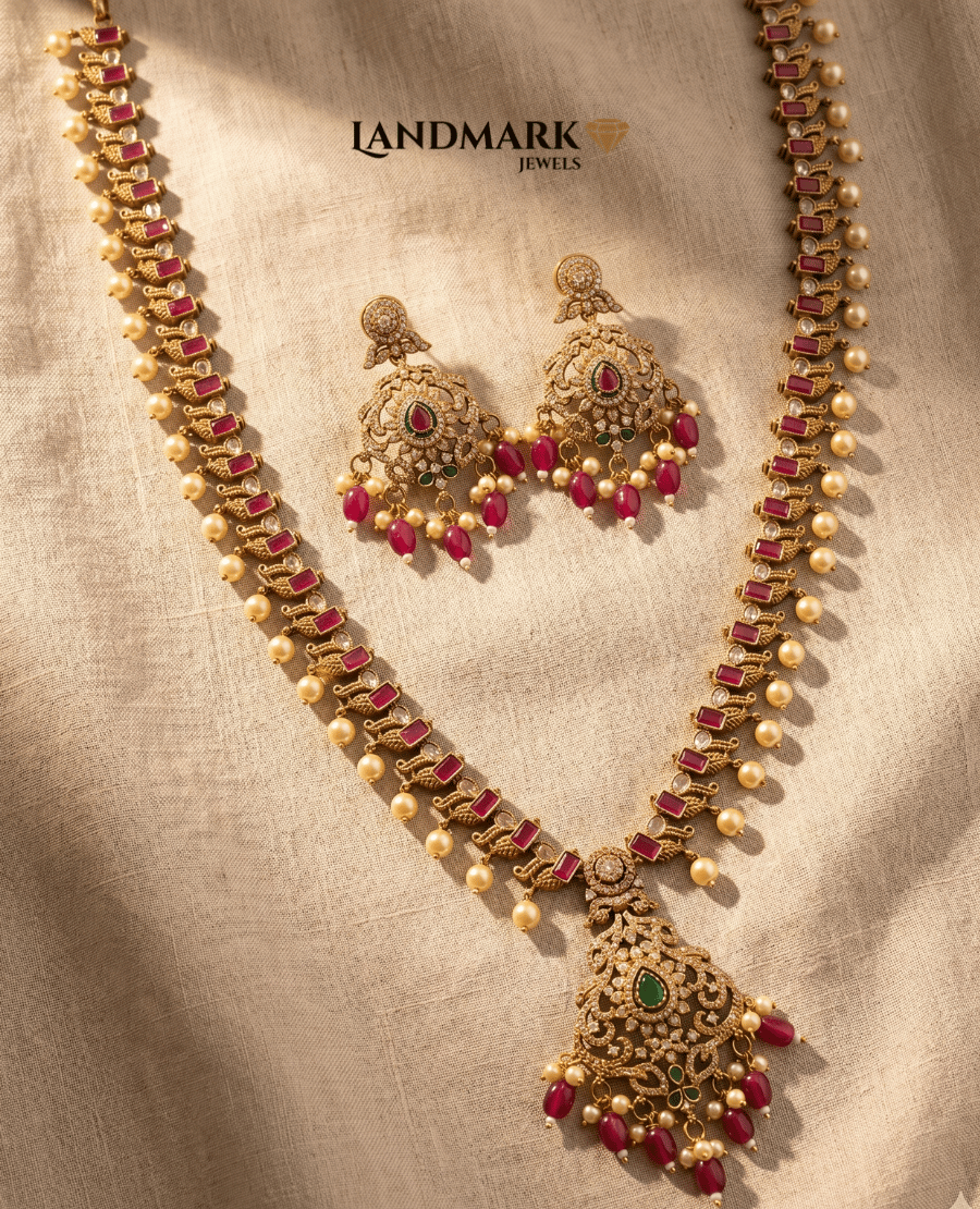 landmark-jewels-antique-gold-plated-long-haram-necklace-and-earring-set-featuring-red-ruby-stones-green-emerald-accents-and-hanging-pearl-drops-on-a-beige-fabric-background