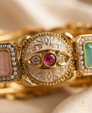 Pair of gold-plated Indian Meenakari bangles with ruby and emerald stones on a beige fabric background.