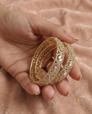 A pair of gold-plated American Diamond bangles featuring circular patterns and central baguette-cut stones, resting on a soft pink velvet surface with a Landmark Jewels brand tag.