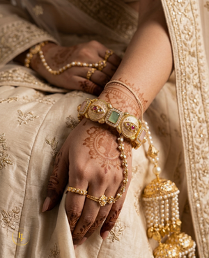 Pair of gold-plated Indian Meenakari bangles with ruby and emerald stones on a beige fabric background.