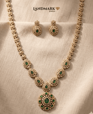 "Landmark Jewels long antique gold necklace set featuring green emerald teardrop stones and white Polki accents with matching earrings on a beige fabric background."