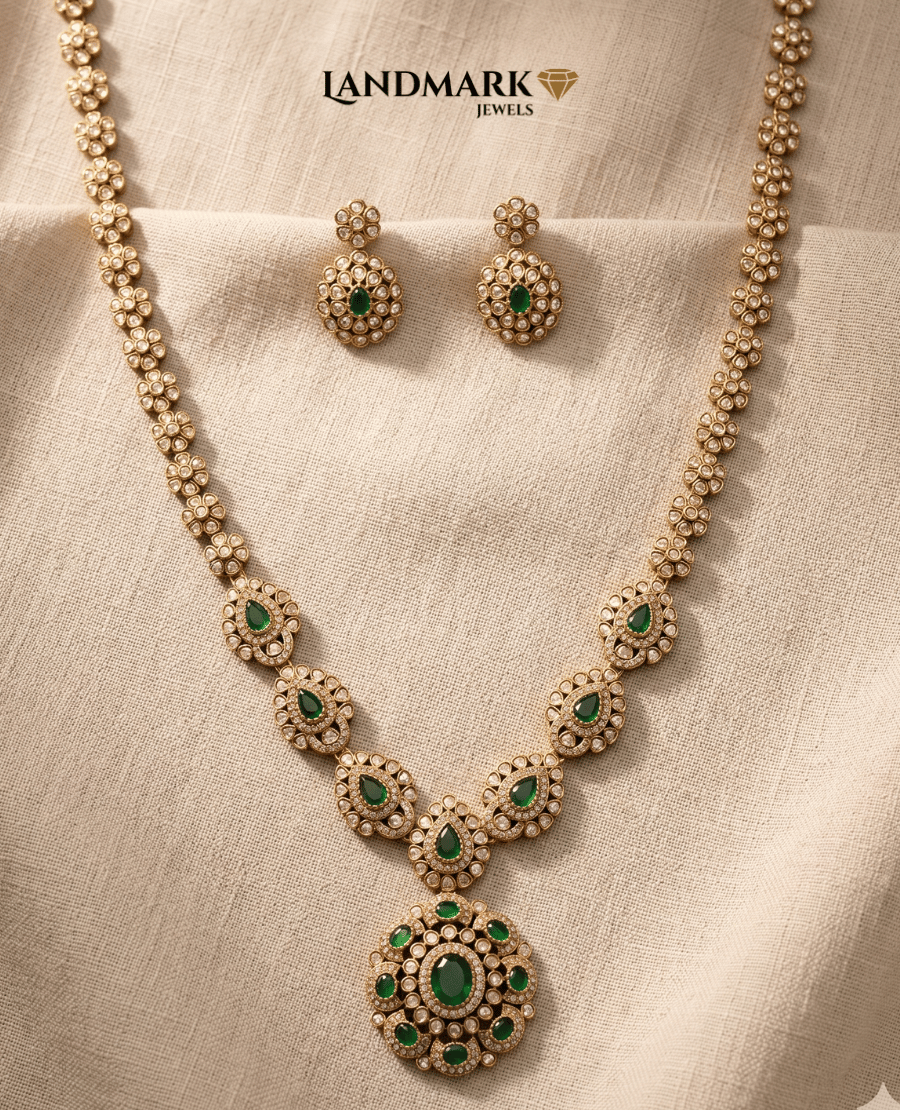 landmark-jewels-long-antique-gold-necklace-set-featuring-green-emerald-teardrop-stones-and-white-polki-accents-with-matching-earrings-on-a-beige-fabric-background