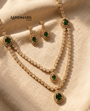 Landmark Jewels imitation gold-toned double-layered necklace and matching drop earrings featuring pear-shaped emerald green stones and white crystal borders on a beige fabric background.