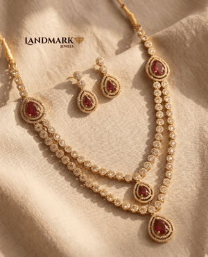 Landmark Jewels gold-plated dual-strand necklace and earring set featuring pear-cut ruby red stones and white CZ accents on a beige fabric background.