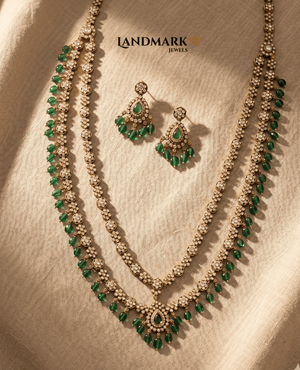 Landmark Jewels antique gold-plated double-layer long necklace and earring set featuring white Polki stones and emerald green bead droplets on a beige fabric background.
