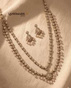 Landmark Jewels antique gold-plated double-layer long necklace set featuring white Polki stones and light pink rose quartz beads with matching drop earrings on a beige fabric background.