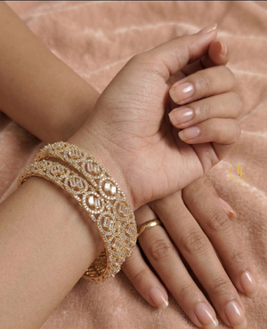 A pair of gold-plated American Diamond bangles featuring circular patterns and central baguette-cut stones, resting on a soft pink velvet surface with a Landmark Jewels brand tag.