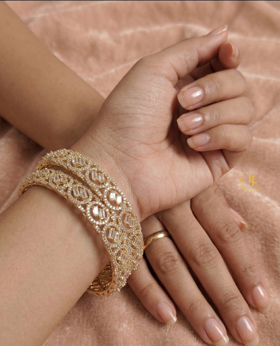 a-pair-of-gold-plated-american-diamond-bangles-featuring-circular-patterns-and-central-baguette-cut-stones-resting-on-a-soft-pink-velvet-surface-with-a-landmark-jewels-brand-tag