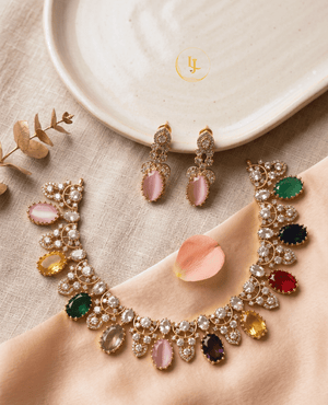 Overhead view of a gold-finish imitation necklace with colorful oval stones and matching earrings displayed on a cream fabric and ceramic plate.