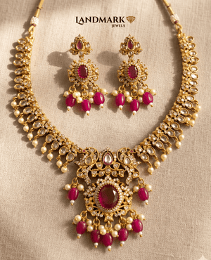Close-up of a Landmark Jewels antique gold-plated necklace and earring set featuring oval rubies, white kundan stones, and hanging pearl drops on a textured beige background.