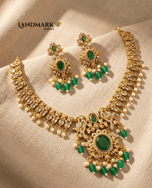 Landmark Jewels antique gold-plated choker necklace and earrings set featuring emerald green stones, white crystals, and hanging pearls on a beige fabric background.