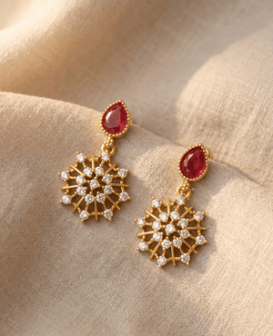 Pair of gold-plated drop earrings from Landmark Jewels featuring a teardrop ruby red stone and a crystalline snowflake drop on a beige fabric background.