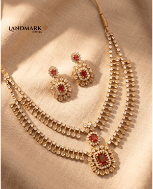 Landmark Jewels two-layer gold-plated necklace set featuring red emerald-cut rubies, white AD stones, and matching drop earrings on a neutral fabric background.