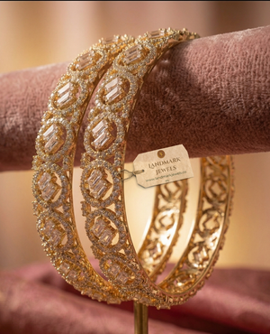 A pair of gold-plated American Diamond bangles featuring circular patterns and central baguette-cut stones, resting on a soft pink velvet surface with a Landmark Jewels brand tag.