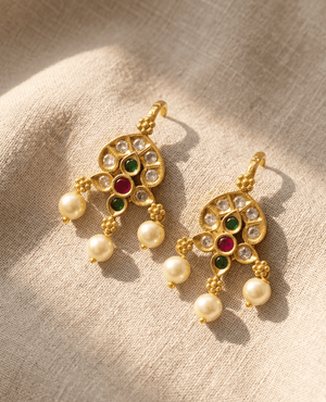 Landmark Jewels traditional matte gold chandbali earrings featuring kundan, ruby red, emerald green stones, and hanging pearls on a beige fabric background.