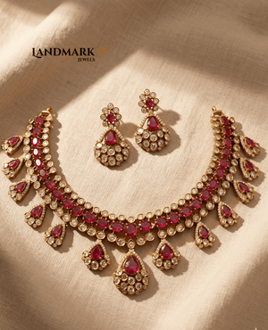 Landmark Jewels gold-plated imitation necklace and earring set featuring oval red rubies and round white crystal accents on a beige fabric background.