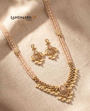 Landmark Jewels imitation jewelry set featuring a three-strand rose quartz and pearl long necklace with a detailed gold-toned floral pendant and matching drop earrings on a beige fabric background.