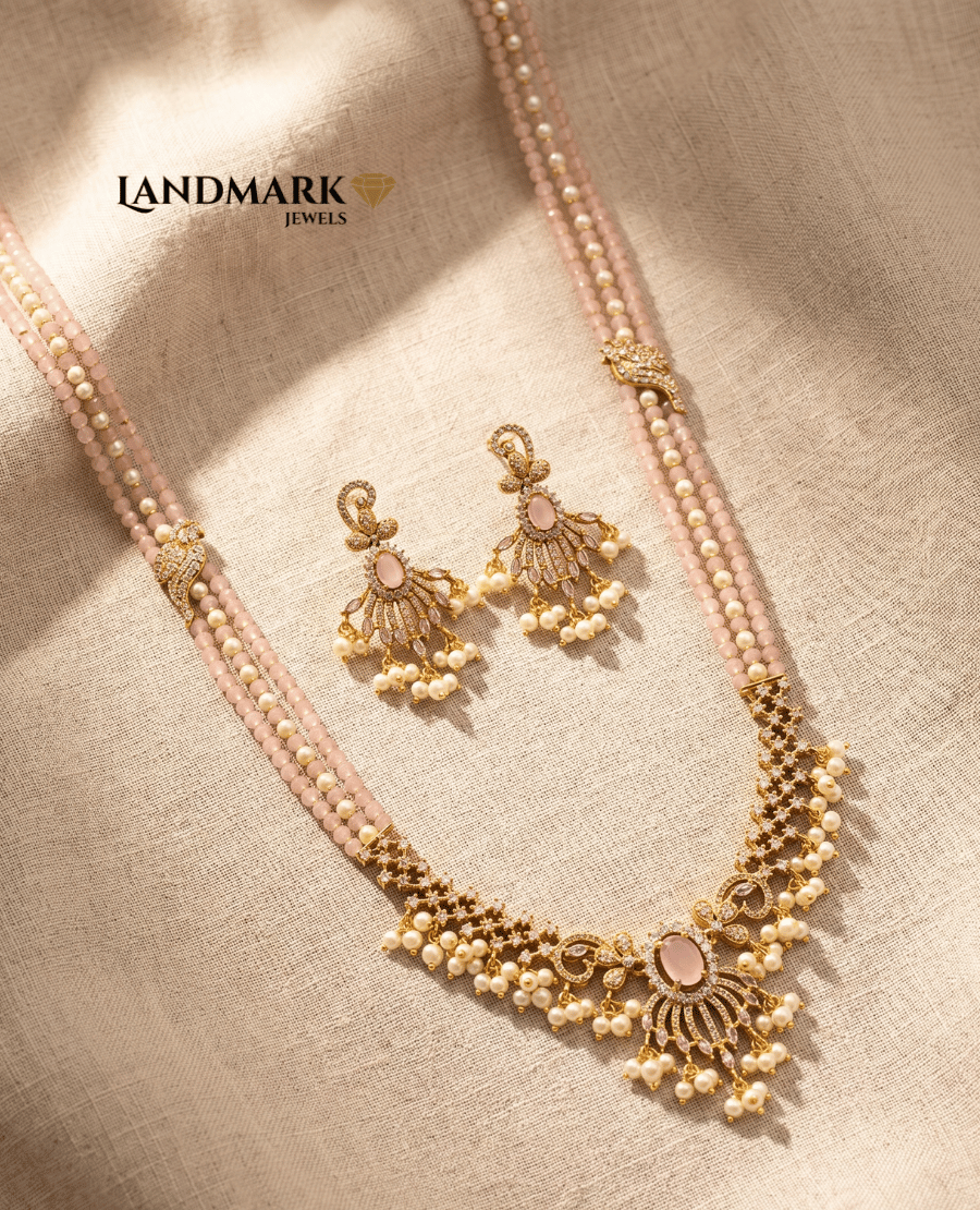 landmark-jewels-imitation-jewelry-set-featuring-a-three-strand-rose-quartz-and-pearl-long-necklace-with-a-detailed-gold-toned-floral-pendant-and-matching-drop-earrings-on-a-beige-fabric-background