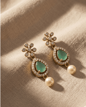 Pair of gold-plated dangler earrings by Landmark Jewels featuring a floral stone stud, a pear-shaped pastel green stone surrounded by crystals, and a hanging white pearl drop on a textured beige fabric background.
