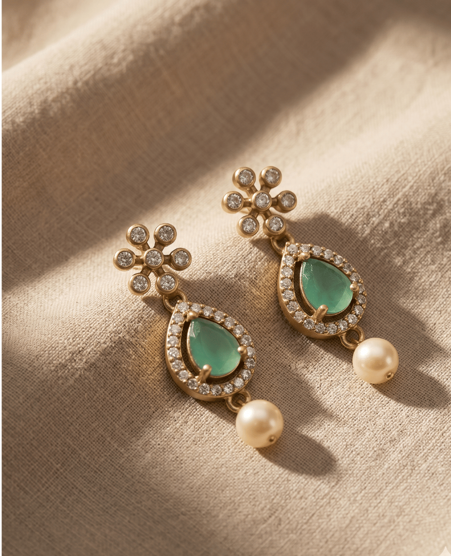 pair-of-gold-plated-dangler-earrings-by-landmark-jewels-featuring-a-floral-stone-stud-a-pear-shaped-pastel-green-stone-surrounded-by-crystals-and-a-hanging-white-pearl-drop-on-a-textured-beige-fabric-background