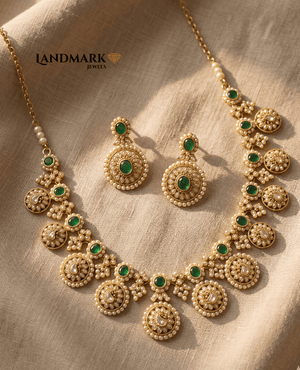 Landmark Jewels antique gold-plated necklace and earring set featuring circular floral motifs, emerald-green stones, and white pearl bead borders on a beige fabric background.