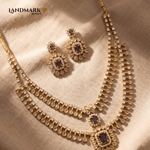 Landmark Jewels double-layered gold-toned necklace and earring set featuring purple emerald-cut stones and teardrop crystal accents on a beige fabric background.