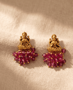 Antique gold temple jewelry earrings featuring a detailed 3D motif of Goddess Lakshmi on a lotus with a dense fringe of faceted maroon ruby-red beads.