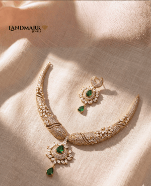 Landmark Jewels gold-plated necklace and earring set featuring emerald-green teardrop stones and intricate white cubic zirconia floral patterns on a neutral fabric background.