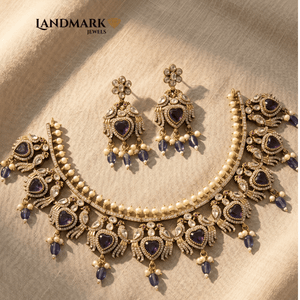 Landmark Jewels imitation jewelry set featuring a gold-toned choker and earrings with purple heart stones, white pearls, and peacock motifs on a beige fabric background.