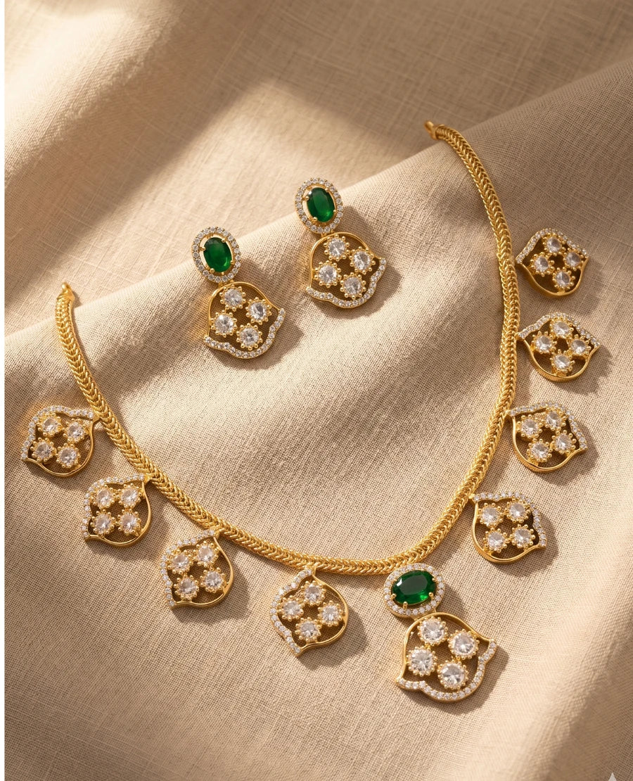 gold-plated-necklace-set-with-emerald-green-stones-and-american-diamond-leaf-pendants-with-matching-earrings-on-beige-fabric