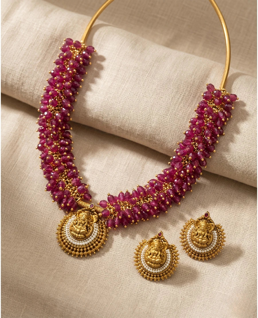 antique-gold-ruby-beaded-hasli-necklace-with-lakshmi-pendant-and-matching-pearl-earrings-on-beige-fabric